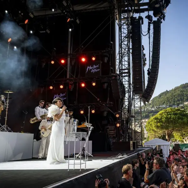 Une musicienne vêtue d'une robe blanche et d'ailes d'ange se produit sur une scène en plein air au festival de jazz de Montreux, avec des membres de l'orchestre, des lumières et une foule en avant, entourée d'arbres et de montagnes en arrière-plan.