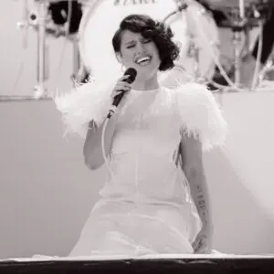 Une femme vêtue d'une robe blanche à plumes s'agenouille sur scène tout en chantant passionnément dans un microphone. Une batterie est visible à l'arrière-plan. L'image est en noir et blanc.