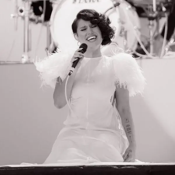 Une femme vêtue d'une robe blanche à plumes s'agenouille sur scène tout en chantant passionnément dans un microphone. Une batterie est visible à l'arrière-plan. L'image est en noir et blanc.