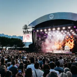 Une foule nombreuse assiste à un concert en plein air au coucher du soleil, avec une scène brillamment éclairée, un écran montrant un artiste, et des montagnes et des arbres en arrière-plan.