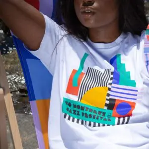A woman sits outdoors in sunlight, wearing the T-Shirt Camille Walala, 2022, featuring a colorful abstract design and "50 Montreux Jazz Festival" text.