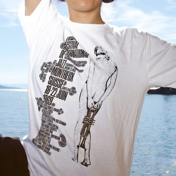 A person wearing a T-Shirt Eric Wondergem, 1969, with black text and graphics for the 1969 Montreux Jazz Festival, stands outdoors by a body of water on a sunny day.