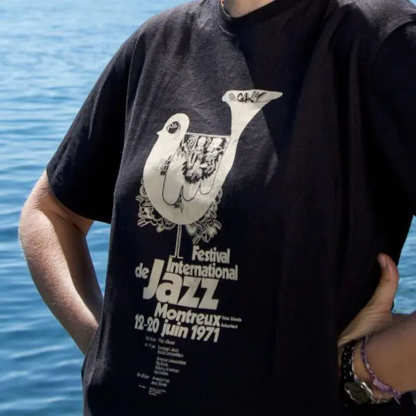 Une personne portant un t-shirt noir avec un graphisme blanc pour le Montreux Jazz Festival de 1971, avec un oiseau stylisé et des détails sur l'événement, debout près d'un plan d'eau.
