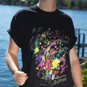 Portant le "T-Shirt Jean Tinguely, 1982" avec un art abstrait coloré et un texte écrit à la main, une personne se tient au bord d'un lac à Montreux sous la lumière du jour, entourée d'eau et de verdure floues en arrière-plan.