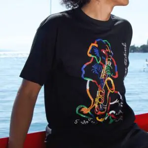 Vêtue d'un T-shirt Rolf Knie, 1996, représentant un saxophoniste abstrait et un texte de festival, une personne est assise au bord de l'eau, sous le soleil.