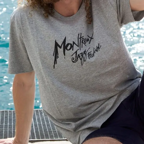 Une personne portant un T-Shirt Montreux Jazz Festival, Gris, est assise au bord de l'eau, son bras et ses jambes étant partiellement visibles. La scène présente un front de mer ensoleillé et animé en arrière-plan.