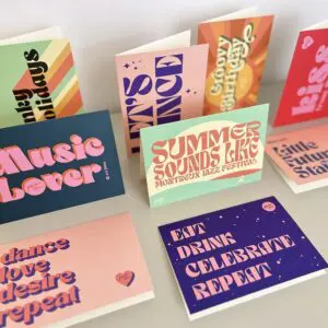 The Dance Love Desire Repeat Card features bold, retro-style typography and vibrant colors, displayed both upright and flat. The card showcases phrases like “Music Lover,” “Dance Love Desire,” and “EAT DRINK CELEBRATE REPEAT.”.
