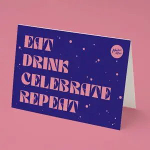 The Eat Drink Celebrate Repeat Card displays bold pink text on a dark blue background reading: EAT DRINK CELEBRATE MUSIC REPEAT, set on a pink surface with scattered dots and a small circular logo in the top corner.