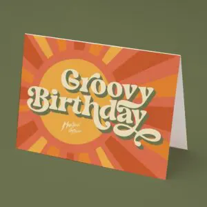 The Groovy Birthday Card features retro, wavy "Groovy Birthday" text in cream on an orange and yellow sunburst background, inspired by the lively Montreux Festival. The card is pictured standing on an olive green surface.