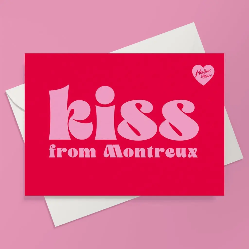 The Kiss, Carte de vœux est une carte postale rouge avec un texte rose en gras "Kiss from Montreux", un petit cœur blanc en haut à droite indiquant "Montreux vibes", inspirée par le festival de musique Kiss Montreux, présentée sur une enveloppe blanche sur fond rose.