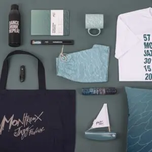 A flat lay photo displays Montreux Jazz Festival merchandise on a green background, including a T-Shirt Guillaume "SupaKitch" Grando 2023, tote bag, notebook, mug, water bottle, swim shorts, pens, pillow, mini sailboat, and lighter.