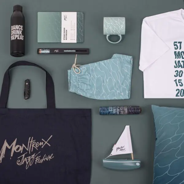 A flat lay photo displays Montreux Jazz Festival merchandise on a green background, including a T-Shirt Guillaume "SupaKitch" Grando 2023, tote bag, notebook, mug, water bottle, swim shorts, pens, pillow, mini sailboat, and lighter.