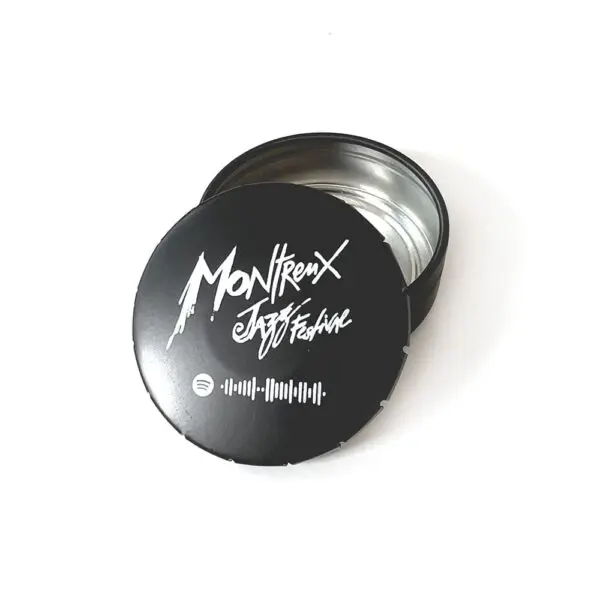 Une boîte ronde noire avec un couvercle partiellement enlevé affichant le Montreux Jazz Festival en écriture blanche et un code Spotify. L'intérieur de la boîte est vide et métallique.