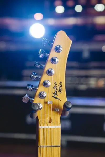 Close-up of a Detroit, Guitar Collection, Limited Series headstock with tuning pegs and signature, set against blurred stage lights, capturing the vibrant energy of the city's iconic music scene.