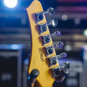 Close-up of the back of a guitar headstock from the Detroit Guitar Collection, Limited Series, featuring engraved "DETROIT" tuning pegs, inspired by the Montreux Jazz Guitar style, with blurred stage lights in the background.