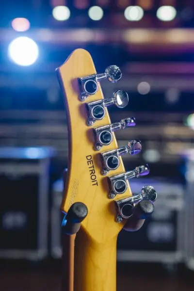 Close-up of the back of a guitar headstock from the Detroit Guitar Collection, Limited Series, featuring engraved "DETROIT" tuning pegs, inspired by the Montreux Jazz Guitar style, with blurred stage lights in the background.