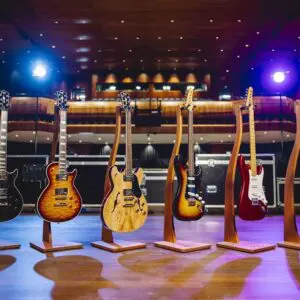 Six Montreux Guitar Collection Limited Series electric guitars are displayed on wooden stands on a concert hall stage under colorful spotlights, with empty seats beyond.