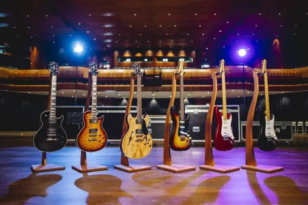 Six Montreux Guitar Collection Limited Series electric guitars are displayed on wooden stands on a concert hall stage under colorful spotlights, with empty seats beyond.