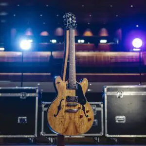 Une guitare électrique semi-hollow Montreux Série limitée de la Collection Guitare, avec une finition en bois naturel, se tient debout sur la scène, éclairée par des lumières bleues et blanches et flanquée de grandes mallettes noires.