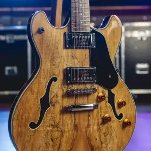 Gros plan d'une guitare électrique semi-hollow avec une finition en bois naturel, un pickguard noir et des boutons de commande dorés, posée sur un support, avec des lumières de scène et des caisses de matériel en arrière-plan flou.