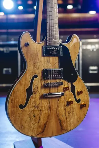 Gros plan d'une guitare électrique semi-hollow avec une finition en bois naturel, un pickguard noir et des boutons de commande dorés, posée sur un support, avec des lumières de scène et des caisses de matériel en arrière-plan flou.