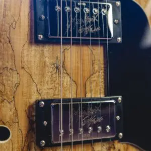 Gros plan du corps d'une guitare électrique, montrant le grain du bois, les micros en métal avec des signatures gravées, les cordes, un pickguard noir, et une partie d'un chevalet argenté et d'un bouton de tonalité ambré.