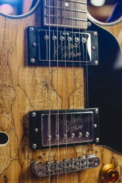 Gros plan du corps d'une guitare électrique, montrant le grain du bois, les micros en métal avec des signatures gravées, les cordes, un pickguard noir, et une partie d'un chevalet argenté et d'un bouton de tonalité ambré.