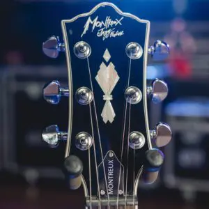 Gros plan d'une tête de guitare montrant le logo Montreux, des chevilles et un motif décoratif incrusté, avec un arrière-plan flou montrant l'éclairage de la scène.
