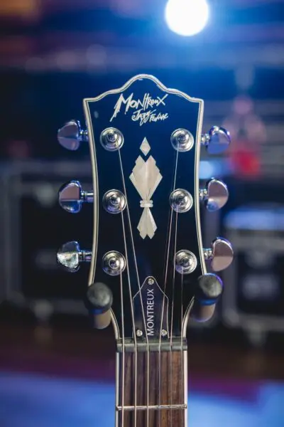 Gros plan d'une tête de guitare montrant le logo Montreux, des chevilles et un motif décoratif incrusté, avec un arrière-plan flou montrant l'éclairage de la scène.