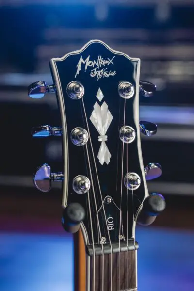 Gros plan d'une tête de guitare avec des chevilles d'accordage, avec un motif incrusté et le logo "Montreux Jazz Festival" dans la partie supérieure. L'arrière-plan est flou et éclairé en bleu et en violet.