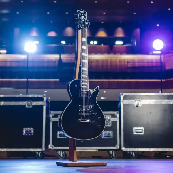 Centrée sur la scène, la Tokyo, Guitar Collection, Série limitée - une guitare électrique noire sur un support en bois - est mise en lumière parmi des caisses de matériel, évoquant l'ambiance d'une salle de concert de Tokyo.