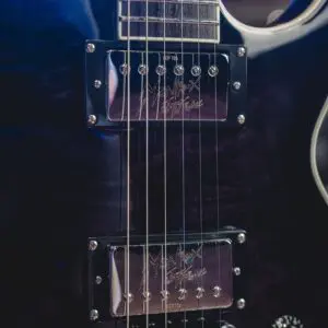 Gros plan d'une guitare électrique montrant deux micros, des cordes en métal, une partie du chevalet, un bouton de commande et des signatures gravées sur les micros. Le corps de la guitare a une finition brillante et sombre.