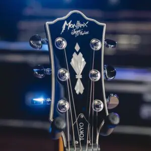 Gros plan sur la tête d'une guitare noire avec des chevilles argentées, portant les inscriptions "Montreux Jazz Festival" et "TOKYO". Les lumières vives de la scène et l'arrière-plan flou suggèrent un spectacle.