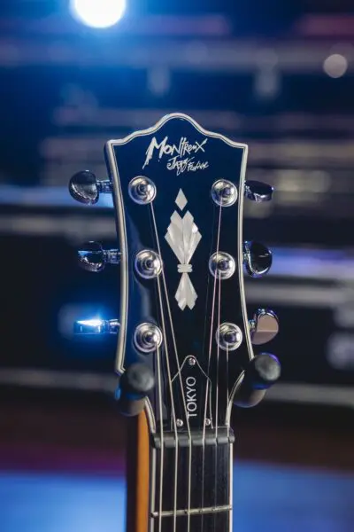 Gros plan sur la tête d'une guitare noire avec des chevilles argentées, portant les inscriptions "Montreux Jazz Festival" et "TOKYO". Les lumières vives de la scène et l'arrière-plan flou suggèrent un spectacle.