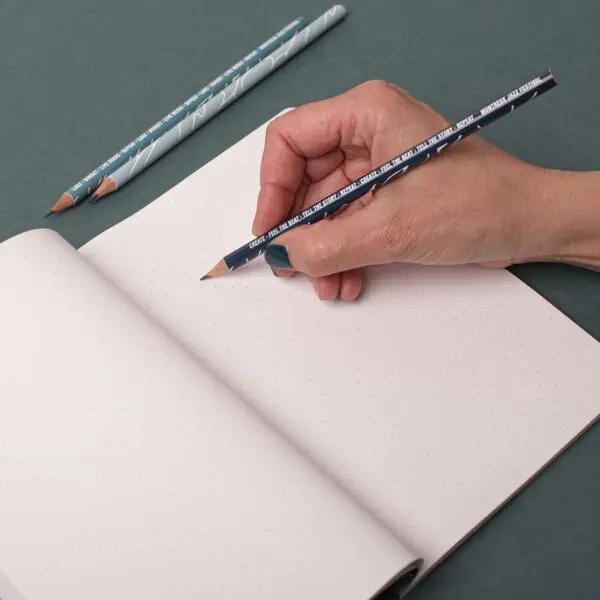 A hand holds a pencil over an open Stone Paper Notebook, Guillaume “SupaKitch”, ready to write, while another pencil rests on the green surface beside the notebook.