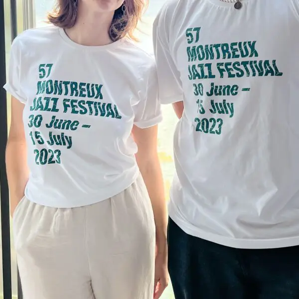 Deux personnes se tiennent côte à côte et portent des t-shirts blancs assortis aux motifs de Guillaume "SupaKitch" Grando, 2023, avec un lettrage texturé vert portant la mention "57 Montreux Jazz Festival, 30 juin - 15 juillet 2023". Leurs visages sont partiellement hors champ.