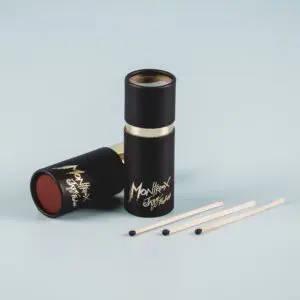 Deux boîtes d'allumettes cylindriques noires avec des lettres dorées, dont l'une est ouverte pour révéler une surface de frappe rouge, reposent sur un fond bleu clair à côté de trois allumettes en bois avec des pointes noires.