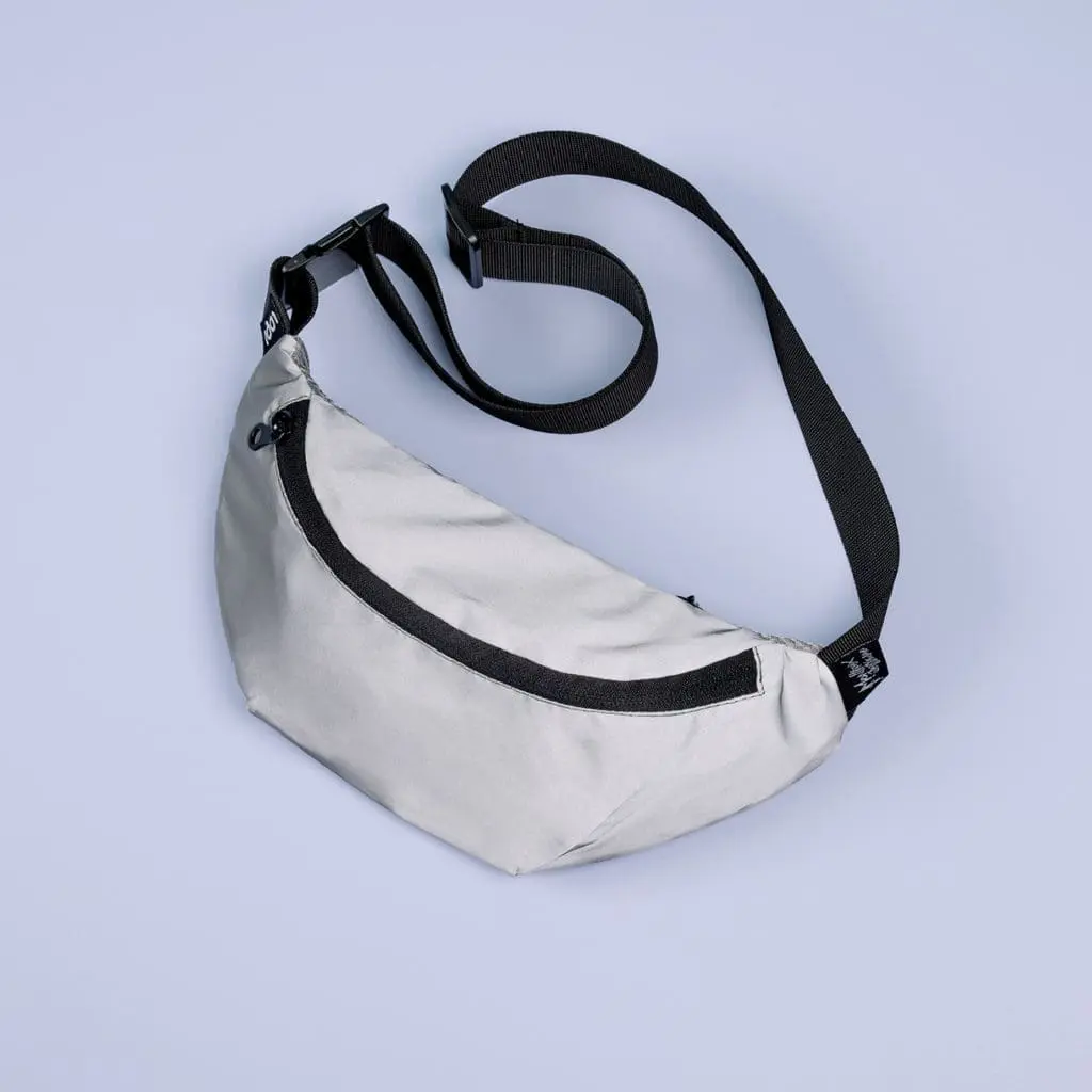 Un Sac banane mini réfléchissante gris clair avec une sangle réglable noire et une fermeture éclair noire est présenté sur un fond bleu pâle.