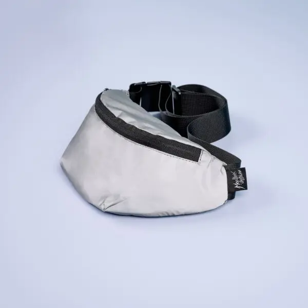 The Reflective Mini Bumbag in light gray, featuring a black zipper, adjustable strap, and small black tag, is displayed on a smooth light blue surface.