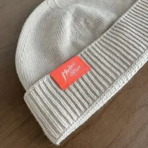 A beige knit beanie called the Beige Hat, Montreux Jazz Festival, features a classic Cousteau style with a folded brim and an orange rectangular tag with white handwritten-style text, shown resting on a wooden surface.