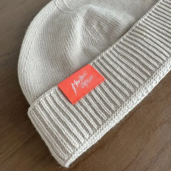 A beige knit beanie called the Beige Hat, Montreux Jazz Festival, features a classic Cousteau style with a folded brim and an orange rectangular tag with white handwritten-style text, shown resting on a wooden surface.