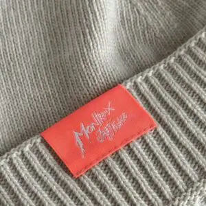 A close-up of the Beige Hat from Montreux Jazz Festival, a knitted Cousteau-style beanie with a red rectangular tag featuring white stylized festival text.