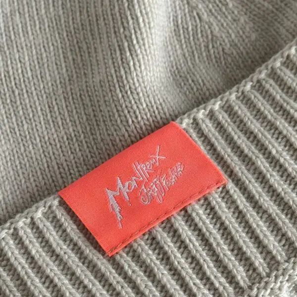A close-up of the Beige Hat from Montreux Jazz Festival, a knitted Cousteau-style beanie with a red rectangular tag featuring white stylized festival text.