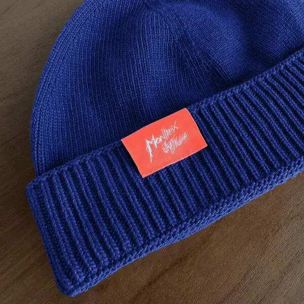 The Blue Hat, Montreux Jazz Festival is a knit beanie with a folded brim and a red patch featuring "Montreux Jazz Festival" in white handwritten text. Its classic Cousteau-inspired style adds flair to any look. Displayed on a wooden surface.