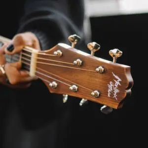 A person with black nail polish holds the neck of a Miami Electro Acoustic Guitar, Limited Series, focusing on the tuning pegs and headstock, with the Miami branding visible against a dark, blurred background.