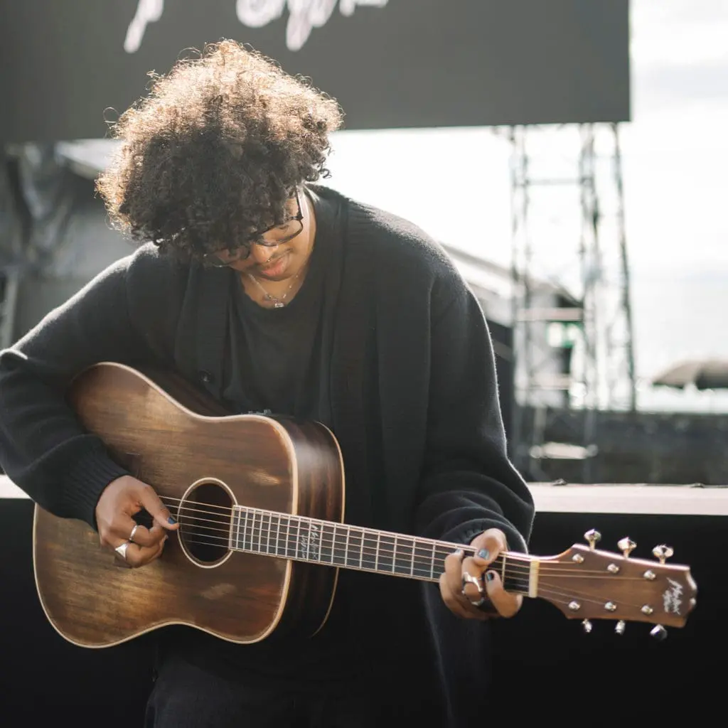Une personne aux cheveux frisés et aux lunettes joue du Miami, Guitare électro-acoustique, Série limitée en plein air au Montreux Jazz. Elle porte un pull sombre et des bagues ; à l'arrière-plan se trouvent une installation de scène floue et une grande enseigne.