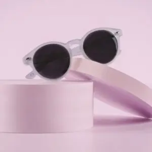 Fly Bruxelles Cristal Glasses, featuring white frames and round black lenses inspired by Montreux Jazz style, are showcased on pastel pink cylindrical, slanted platforms against a minimalist pink background.