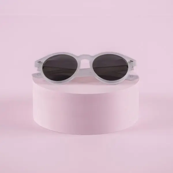 Fly Bruxelles Cristal Glasses, white round sunglasses with dark lenses, rest on a cylindrical pink pedestal against a pastel pink background.