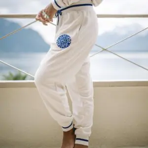 Une personne se tient pieds nus sur un balcon en Pantalon tissu éponge, un pantalon de jogging blanc vintage avec des bordures bleues et un logo circulaire. Un lac pittoresque et des montagnes en arrière-plan évoquent une ambiance de festival vintage.