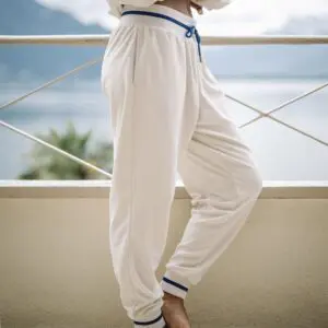 A person stands barefoot on a balcony in Vintage Trousers Terry Cloth with cream color and blue trim. The blurred background highlights water, mountains, and greenery.